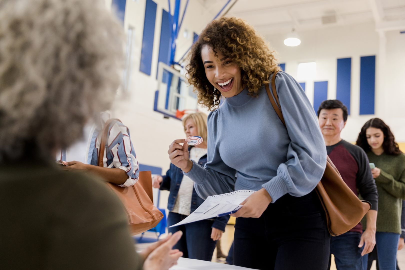 A woman walking up to a person sitting at a desk, the woman is smiling and holding up her "I voted" sticker