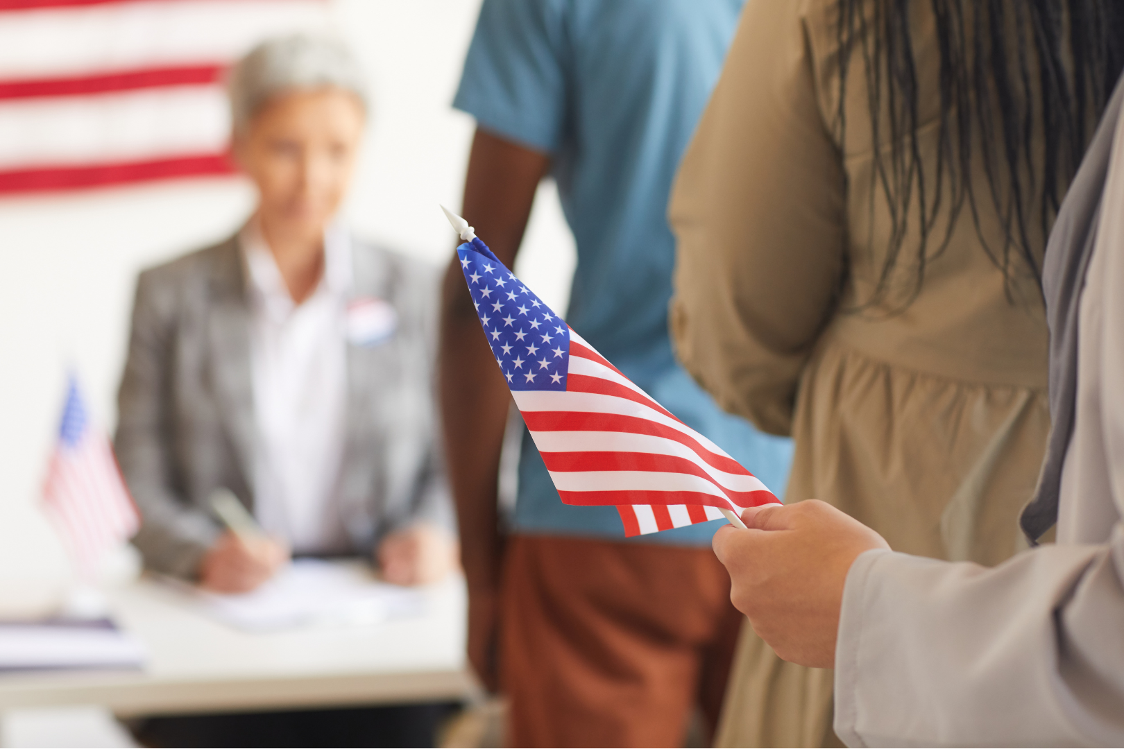 A line of people, one holding a small America flag, waiting to reach a woman sitting at a desk